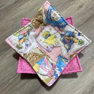 May include: A pink and white quilted bowl cozy with a patchwork design featuring a variety of cute animal and floral motifs. The cozy is made of fabric with a pink polka dot pattern on the back.