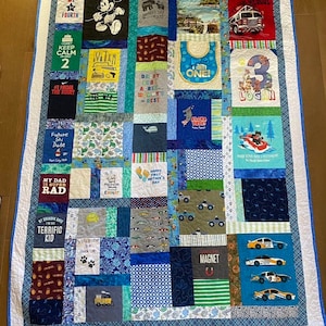 May include: A blue and white patchwork quilt made from t-shirts. The quilt features a variety of designs, including a Mickey Mouse character, a fire truck, a baby bib, and a number 3. The quilt is surrounded by a blue border.