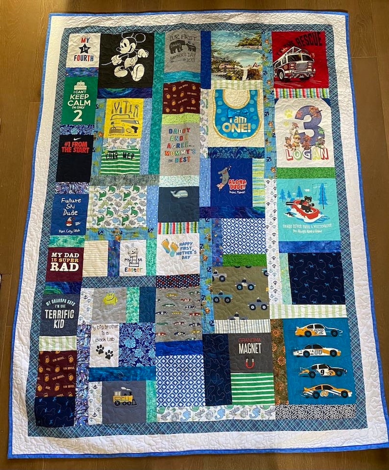 May include: A blue and white patchwork quilt made from t-shirts. The quilt features a variety of designs, including a Mickey Mouse character, a fire truck, a baby bib, and a number 3. The quilt is surrounded by a blue border.