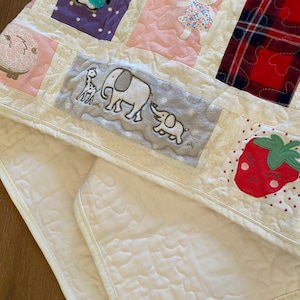 May include: A white quilted baby blanket with a variety of colorful appliques, including a penguin, a bear, elephants, and a strawberry. The blanket is made of soft, comfortable fabric and is perfect for cuddling.