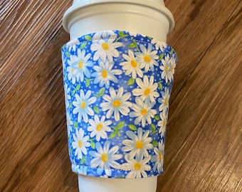 May include: A white coffee cup with a blue and white floral fabric sleeve. The sleeve has a pattern of white daisies with yellow centers on a blue background.