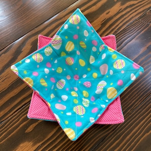 May include: Two handmade bowl cozies. The top cozy is turquoise with a pattern of Easter eggs in yellow, pink, and white. The bottom cozy is pink with white polka dots. Both are square and quilted.