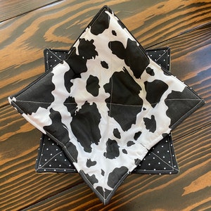 May include: A black and white cow print pot holder with a black and white polka dot lining. The pot holder is folded into a star shape.