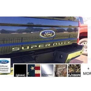 May include: A black Ford Super Duty truck tailgate emblem and lettering set. The Ford logo is oval with blue and silver text. The Super Duty lettering is in a bold font. The image includes swatches for gloss black, American flag, chrome, and Realtree Max-4 camo finishes.