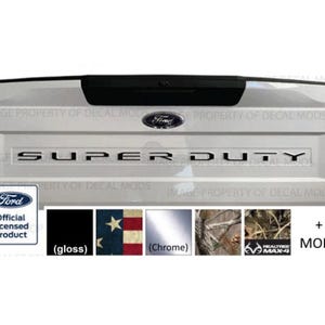May include: A white truck tailgate featuring the Ford logo and the text SUPER DUTY in black lettering. Below, a series of swatches display available finishes including gloss black, American flag pattern, chrome, and Realtree Max-4 camouflage.