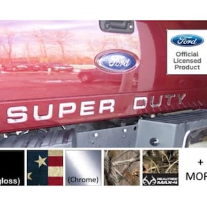 May include: A maroon truck tailgate featuring a chrome Ford emblem and the silver text SUPER DUTY. A white square sticker reads Ford Official Licensed Product. Below are color and pattern swatches including gloss black, American flag, chrome, and Realtree Max-4 camo.