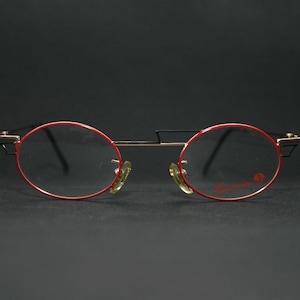 May include: Red and black round eyeglasses with gold accents. The glasses have a thin metal frame and clear lenses.