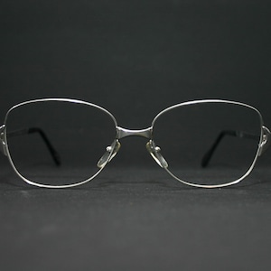 May include: Silver-framed spectacles with a rounded rectangular shape. The spectacles have a thin metal frame and black temples. The lenses are clear, and the nose pads are small and transparent. The background is a solid black.