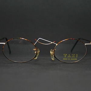 May include: Round, tortoiseshell-framed glasses with gold metal accents. The glasses have clear lenses and the brand name "TAXI MOD. DI VECCHIE" is printed on the right lens.