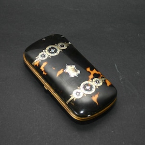 May include: A rectangular black case with a tortoiseshell pattern and gold trim. The case is decorated with ornate silver and gold accents. The design includes circular and star-shaped elements.