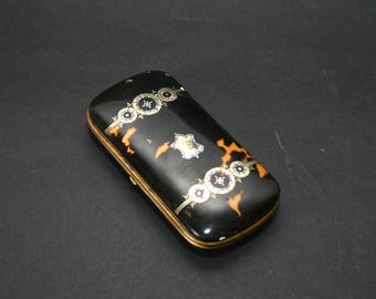 May include: A rectangular black case with a tortoiseshell pattern and gold trim. The case is decorated with ornate silver and gold accents. The design includes circular and star-shaped elements.
