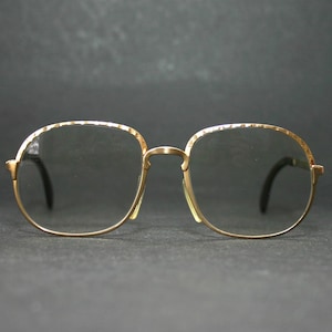 Può includere: Occhiali con montatura dorata con un motivo strutturato sulle montature. Le lenti sono trasparenti e le aste sono nere. Gli occhiali hanno un design classico e vintage.