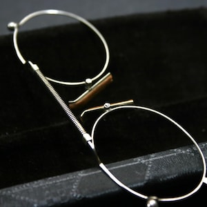 Pode incluir: Óculos sem aro prateados com lentes ovais e dobradiça de mola. Os óculos têm uma pequena ponte nasal redonda e estão colocados sobre um estojo preto forrado a veludo. O design é clássico e elegante.