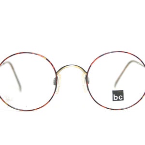 May include: Round tortoiseshell eyeglasses with gold metal accents and clear lenses. The brand name "bc" is printed on the right lens.
