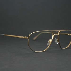 Könnte beinhalten: Goldfarbene Aviator-Brille mit Doppelsteg und klaren Gläsern. Die Fassungen haben ein rechteckiges Design mit einem dezenten Logo auf den Bügeln. Die Bügel sind schwarz. Die Brille ist vor einem dunklen Hintergrund platziert.