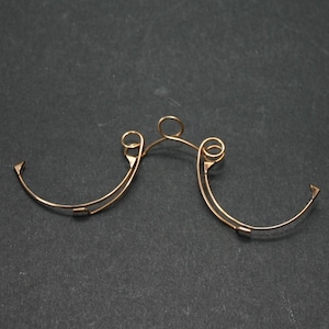 Könnte beinhalten: Ein filigraner, goldfarbener Drahtrahmen, möglicherweise für eine Brille, liegt vor dunklem Hintergrund. Der Rahmen hat gebogene Bügel mit dekorativen Wirbeln und eckigen Spitzen, verbunden durch einen Steg und eine kleine Schlaufe.