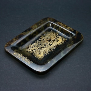 Puede incluir: Una bandeja rectangular con esquinas redondeadas, con un patrón oscuro y moteado. La bandeja está adornada con un diseño de dragón dorado en el centro y detalles decorativos dorados en los bordes. La bandeja está sobre un fondo oscuro.