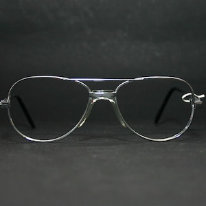 Könnte beinhalten: Silberfarbene Aviator-Brille mit klaren Gläsern. Die Brille hat einen Doppelsteg und gebogene Bügel. Die Gläser sind tropfenförmig. Vor dunklem Hintergrund.