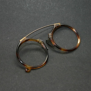 Può includere: Un paio di occhiali rotondi antichi con motivo tartarugato. Gli occhiali hanno un ponte e aste sottili color oro. Le lenti sono assenti e la montatura è appoggiata su una superficie grigio scuro.