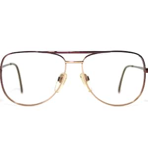 May include: Aviator-style spectacles with a brown and gold metal frame. The lenses are clear, and the nose pads are beige. The glasses have a double bridge and a classic, vintage design.