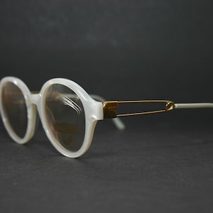 May include: White oval-shaped spectacles with gold-coloured accents. The frames are pearlescent white, and the temples are light grey. The bridge features a gold-coloured safety pin design. The lenses are tinted.