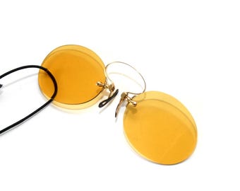 Könnte beinhalten: Randlose Sonnenbrille mit runden, gelb getönten Gläsern. Die Fassungen sind goldfarben, mit einer schwarzen Kordel befestigt. Die Sonnenbrille ist vor einem weißen Hintergrund platziert.