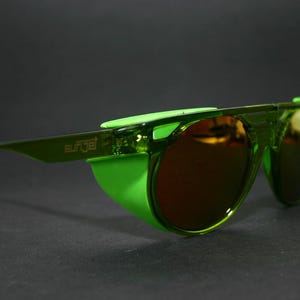 May include: Green sunglasses with a wraparound design. The lenses are a dark amber colour, and the frame and side shields are a bright, translucent green. The word "surge" is printed on the side of the frame.