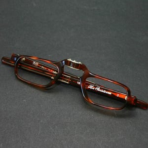 May include: Folded eyeglasses with a tortoiseshell pattern. The glasses have a rectangular frame and the text "La Parisienne" is visible on the arm. The number "3" is also visible on the arm.