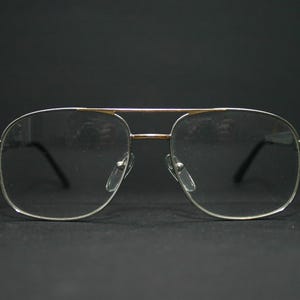 May include: Silver-framed aviator spectacles with clear lenses. The spectacles have a double bridge and black arms. The lenses are rectangular with rounded corners. The spectacles are set against a black background.