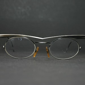 May include: Silver-framed eyeglasses with oval lenses and a distinctive brow bar. The glasses have a minimalist design with a dark bridge and orange nose pads, set against a dark background.
