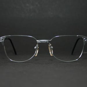Könnte beinhalten: Brille mit silbernem Rahmen und dunklen Gläsern. Die Brille hat ein klassisches Design mit rechteckiger Form und Nasensteg. Die Bügel sind dünn und schwarz. Der Hintergrund ist einfarbig schwarz.