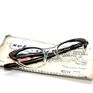 Puede incluir: Gafas vintage de ojo de gato con montura transparente y detalles en negro y dorado, sobre un estuche de papel. El estuche tiene texto que incluye "Selecta" y "Frame France". Las gafas tienen un puente de 42/22.