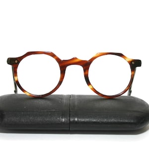 Puede incluir: Gafas redondas con un patrón de carey, apoyadas sobre un estuche negro. Las monturas tienen una forma distintiva con un puente de cerradura. Las lentes son transparentes y las patillas son finas y oscuras.