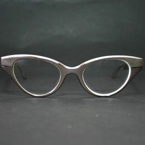 Puede incluir: Un par de gafas con montura plateada con forma de ojo de gato. Las lentes son transparentes y las monturas tienen un acabado mate. Las gafas están sobre un fondo oscuro, resaltando su diseño retro.