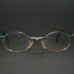 May include: Oval-shaped eyeglasses with a gold-colored frame, blue accents, and clear lenses. The temples are black, and the nose pads are yellow. The brand name is printed in red on the right lens. The glasses are set against a dark background.