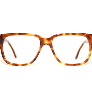 以下が含まれることがあります： べっ甲柄のスクエアフレームメガネ。フレームは温かみのある茶色で、濃い茶色のアクセントが付いています。レンズはクリアで、テンプルも同じ茶色です。メガネはクラシックでレトロなスタイルです。