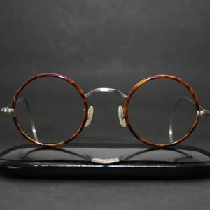 May include: Round eyeglasses featuring a brown and black tortoiseshell pattern on the circular frames, paired with thin silver metal temples and a bridge. The glasses are positioned directly in front of a black, hard-shell protective case.