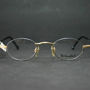 Puede incluir: Gafas de forma ovalada con puente y detalles dorados, monturas plateadas y varillas negras. La marca "Brendel" está escrita en la lente derecha. Las gafas están sobre un fondo oscuro.