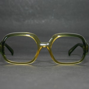 Puede incluir: Un par de gafas con una montura de color verde oliva translúcido con forma rectangular redondeada. La montura tiene un acabado liso y brillante e incluye patillas de color verde oscuro que se extienden desde los lados de los aros.