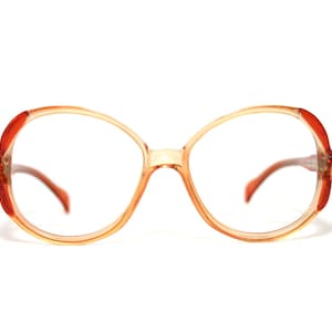 May include: Vintage eyeglasses with a large, round frame. The frame is a light tan color with red accents on the temples. The lenses are clear. A retro accessory.