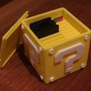 May include: A yellow 3D printed box shaped like a question mark block from the Super Mario Bros. video game. The box has a hinged lid and is designed to hold multiple game cartridges.