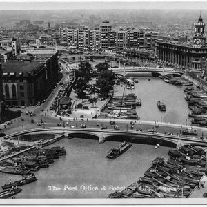May include: A black and white vintage postcard showing a view of the Post Office and Soochow Creek in Shanghai, China. The postcard features a bridge over the creek, with boats docked on either side. The Post Office building is visible in the background.