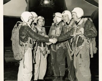 May include: A group of five men in military uniforms and helmets stand in front of a military aircraft. They are all wearing parachutes and are holding onto each other's hands. The aircraft is a bomber with a large, round fuselage and a long, narrow wingspan.
