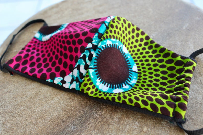 May include: A reusable face mask with a colorful geometric pattern. The mask is made of fabric and has a black elastic band. The pattern features pink, green, blue, white, and brown colors.