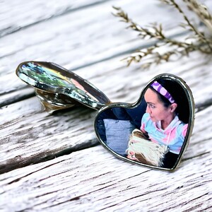 May include: A heart-shaped metal pin featuring a photographic print of a person wearing a purple headband and a light blue and pink hooded sweatshirt. The pin has a silver-toned metallic border and a butterfly clutch backing mechanism visible.