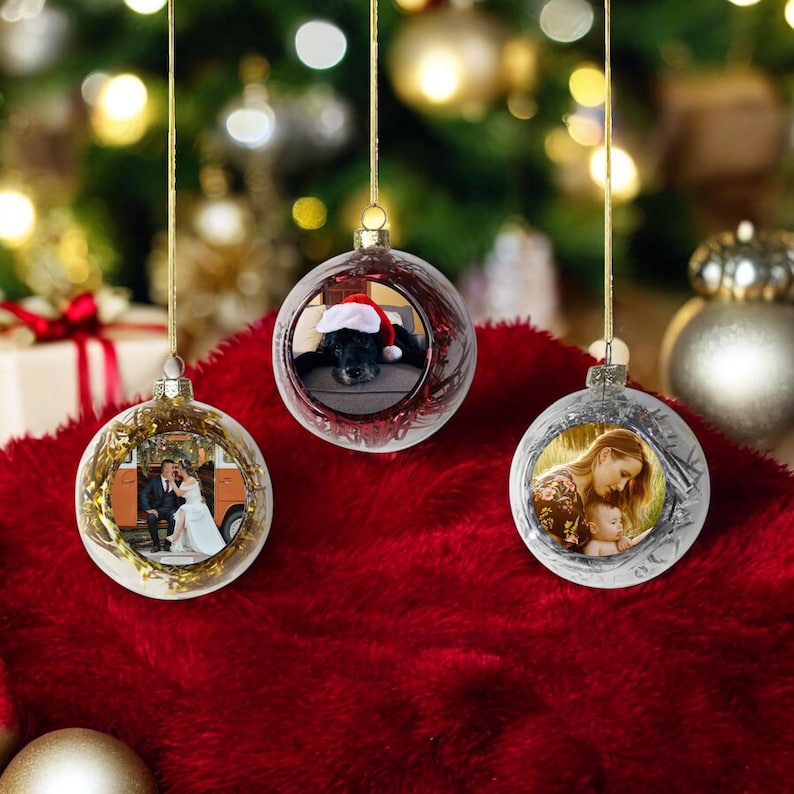 Op de afbeelding: Drie doorzichtige, bolvormige kerstballen die aan gouden koordjes hangen. Elke kerstbal bevat een ronde foto: een koppel, een hond met een kerstmuts en een persoon die een baby vasthoudt. De kerstballen zijn voorzien van metalen accenten.