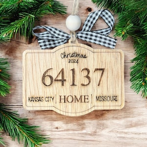 Op de afbeelding: Een houten kerstversiering met een zwart-wit geruit lint, de tekst "Christmas 2024", het nummer "64137" en de tekst "Kansas City HOME Missouri".