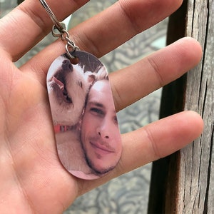 May include: A silver keychain with a photo of a person and a white dog. The photo is printed on a rectangular tag.