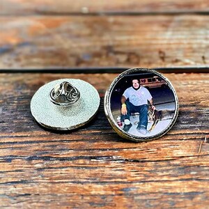 May include: A circular metal pin featuring a photographic print of a person wearing a light-colored t-shirt and dark pants sitting outdoors with a dog. The back of the pin shows a silver-toned textured surface with a metal butterfly clutch fastener.