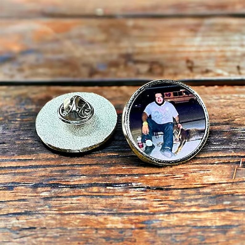 May include: A circular metal pin featuring a photographic print of a person wearing a light-colored t-shirt and dark pants sitting outdoors with a dog. The back of the pin shows a silver-toned textured surface with a metal butterfly clutch fastener.